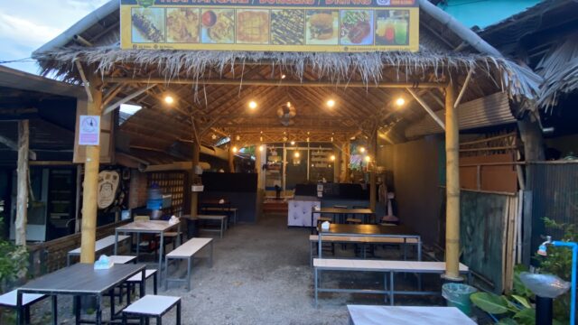 Outdoor Thai food stall with thatched roof, menu signs, and empty tables set on gravel under evening sky.