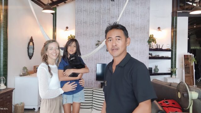 Three people stand indoors, smiling; a girl in the center holds a black cat. The room is modern, with plants, shelves, and a hammock.