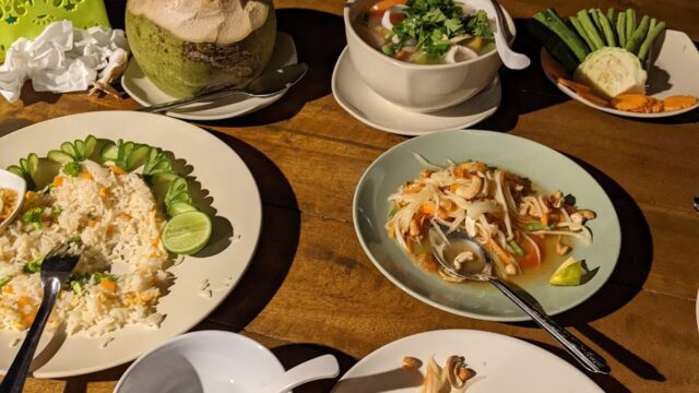 Thai meal on wooden table: rice, coconut drink, soup, salad, fresh veggies, tissues, and utensils; some food partly eaten.