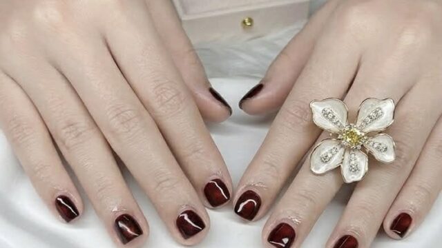 Hands with dark burgundy polished nails, a large flower ring, and a ring box with a diamond ring on a white tray.