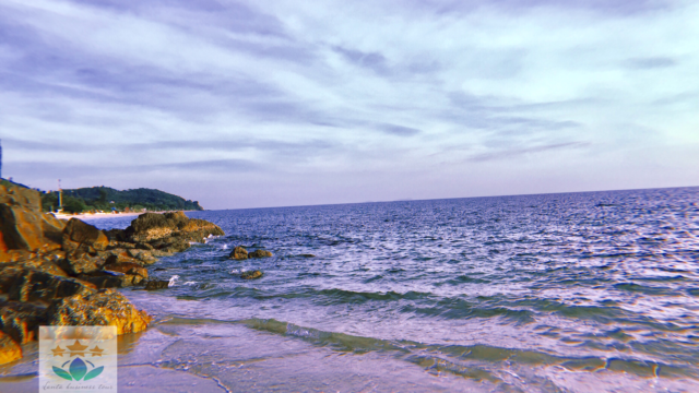 Waves gently crash onto a rocky beach with a distant green hill under a partly cloudy sky at sunset.