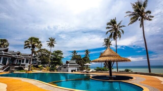 A tropical resort pool with palm trees, a thatched umbrella, and ocean view under a partly cloudy blue sky.