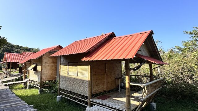 Small bamboo huts with red roofs sit beside a bamboo walkway, surrounded by grass and trees under a clear blue sky.