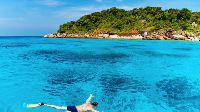 A person snorkeling in clear blue water near a lush green island under a bright blue sky.