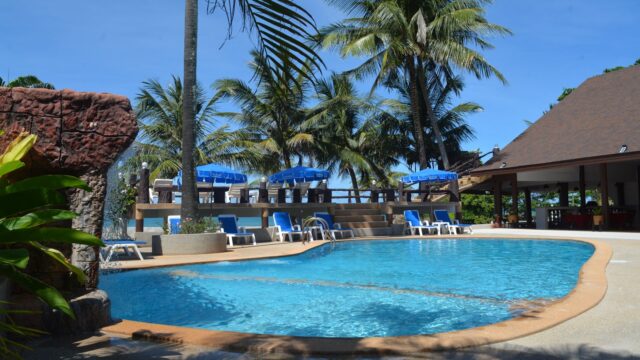 Swimming pool surrounded by palm trees, blue lounge chairs, and umbrellas near a poolside bar on a sunny day.