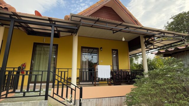 A yellow house with a front porch, green plants, and a partly cloudy sky overhead.
