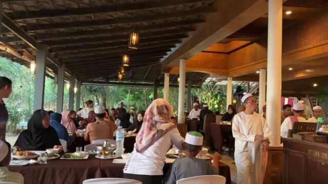 A group of people in traditional attire gather indoors around tables, sharing a meal in a warmly lit setting.