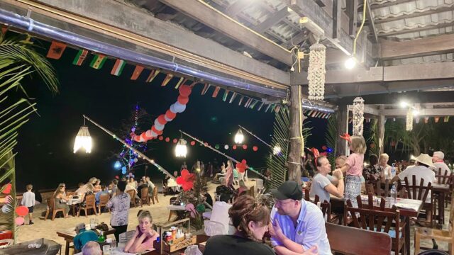 People dining at a beachside restaurant at night, with festive lights, flags, and decorations.