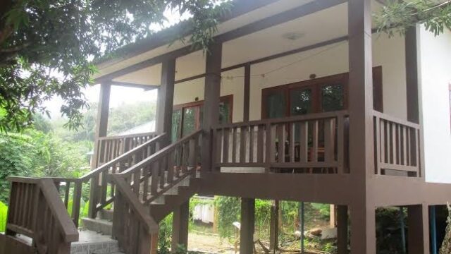 A small house with a wooden porch, brown railings, and stairs, surrounded by trees and greenery.