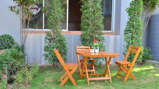 Wooden table with three chairs in a garden, near bushes and a large window on a gray building.