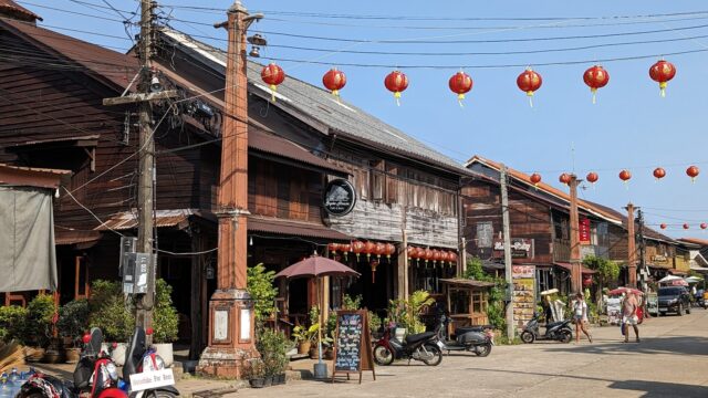 Street with wooden buildings, red lanterns hanging, motorbikes parked, and people walking on a sunny day.