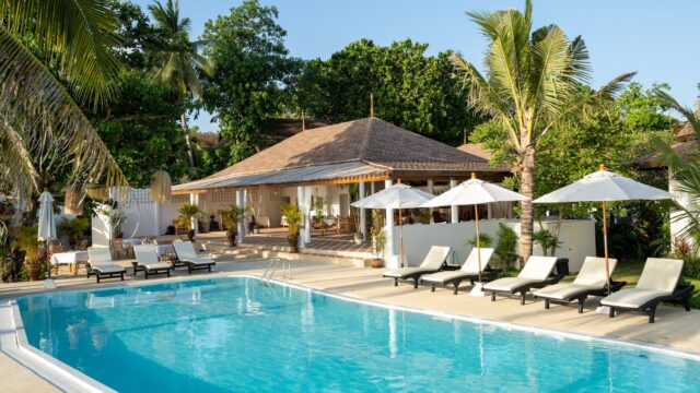 A tropical resort pool surrounded by lounge chairs, umbrellas, palm trees, and a villa in the background under a clear sky.