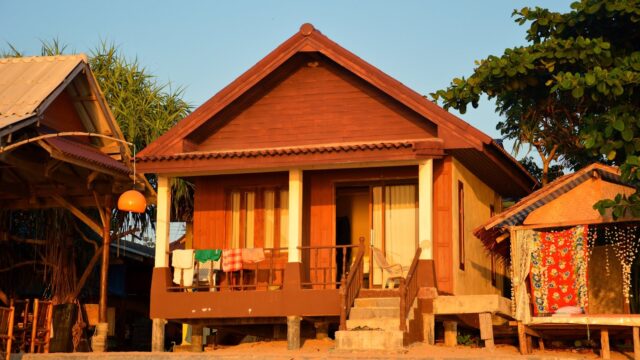 A small wooden beach house with laundry hanging on the porch, lit by warm sunlight, surrounded by trees and sand.