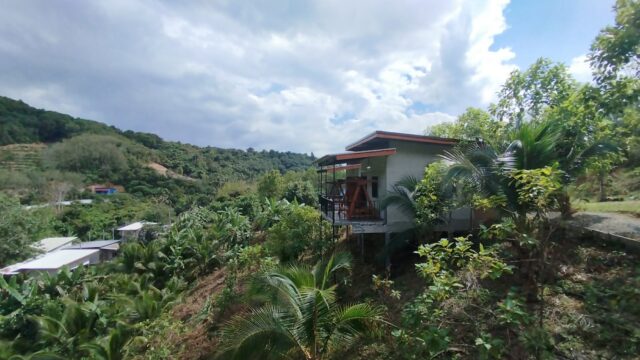 A small house on stilts overlooks lush, green hills and tropical trees under a partly cloudy sky.
