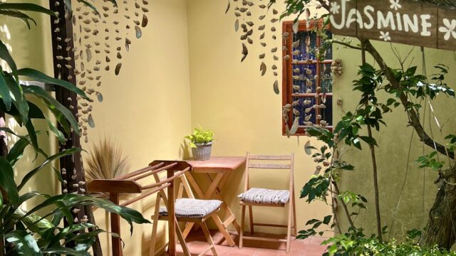 A cozy outdoor patio with two chairs, a small table, plants, and a Jasmine sign, surrounded by greenery and a Welcome mat.