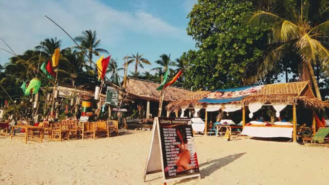 A sandy beach with wooden chairs, a massage sign, palm trees, and colorful flags near thatched-roof huts under a blue sky.
