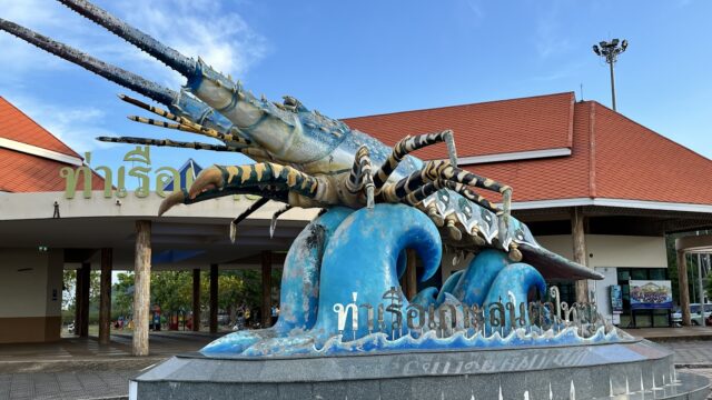 Large blue lobster statue on a blue wave base in front of a building with a red roof and Thai text on the sign.