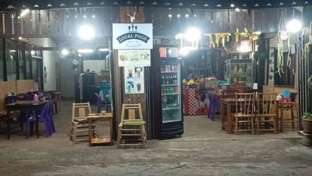 Outdoor local restaurant at night with wooden tables, chairs, a fridge, string lights, and a Local Food sign in the middle.