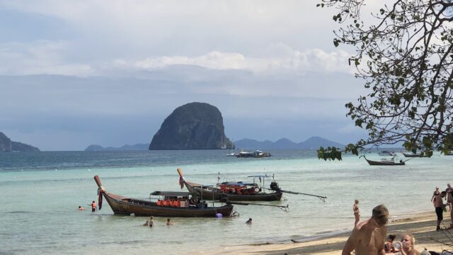 People relax on a sandy beach with boats in the water and a large island rock in the background under a partly cloudy sky.