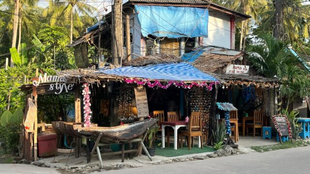 A small, rustic café with outdoor seating, palm trees, and tropical decor sits beside a quiet street under a blue tarp roof.