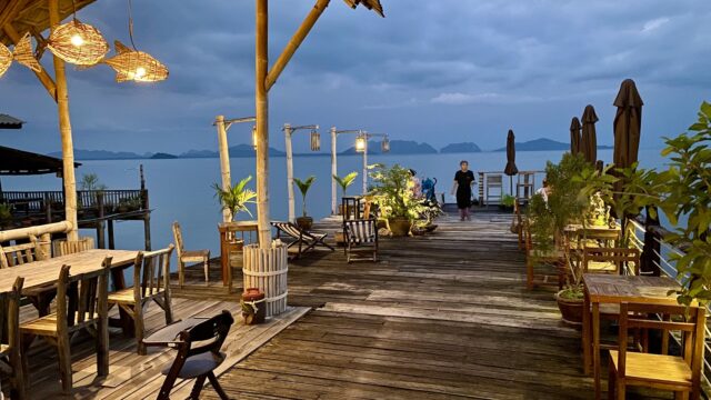 Wooden deck restaurant at dusk with hanging lights, empty tables, plants, and two people walking by the water.