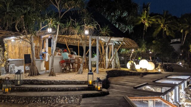 Outdoor beachside bar at night with people sitting, lanterns glowing, and tropical trees surrounding a wooden hut.