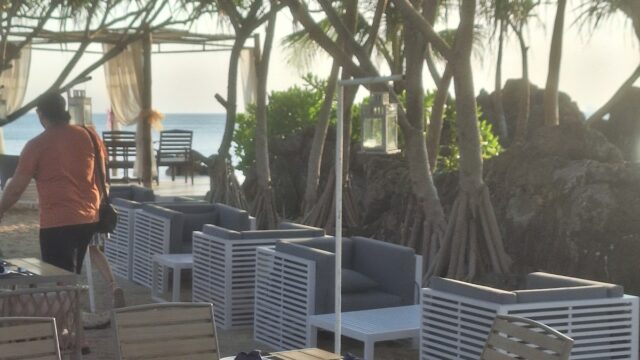 Outdoor beachside dining area with wooden tables and chairs, shaded by trees, with a person walking away.