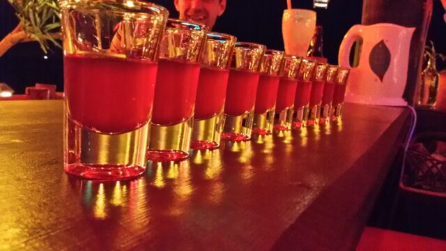 A row of red-colored shots lined up on a bar counter, with people and drinks in the background.