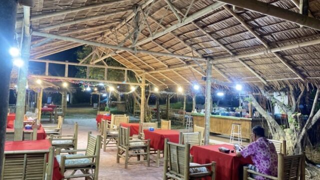 Open-air bamboo restaurant with red tablecloths, wooden chairs, string lights, and one person sitting at a table.