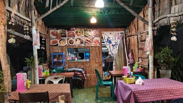 Small rustic outdoor restaurant with bamboo walls, colorful chairs, and food photos hanging above the counter at night.