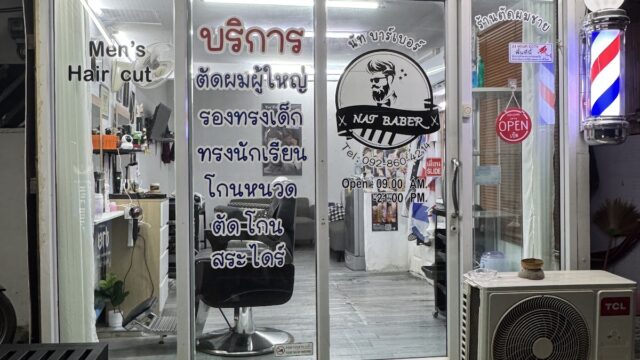 A barber shop entrance with glass doors, service signs in Thai, an open sign, and a traditional barber pole on the right.