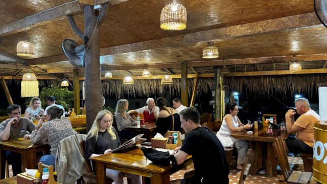 People dining at a rustic, open-air restaurant with wooden tables, warm lighting, and a relaxed atmosphere.