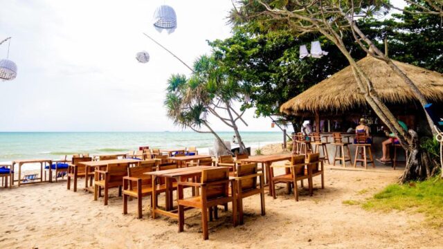 Wooden tables and chairs on a sandy beach, with a thatched-roof bar and the ocean in the background.