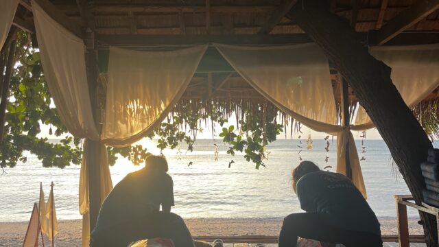Two people receive outdoor massages on mats near a beach at sunset, under a shaded canopy with curtains and trees.