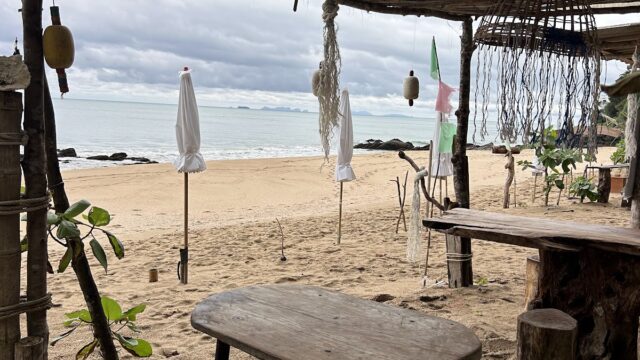 A rustic wooden table and stools under a canopy on a sandy beach, with closed umbrellas and a cloudy sky over the sea.