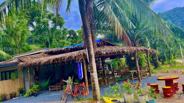 A tropical café with palm trees, hammocks, a red bicycle, and a colorful kayak in a lush, green setting.