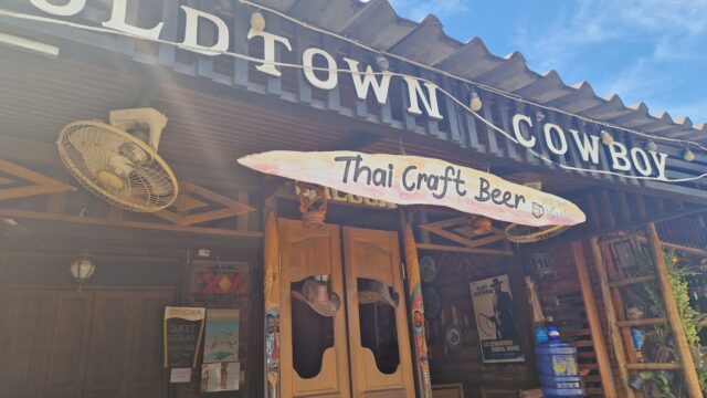 Front view of Old Town Cowboy bar with a Thai Craft Beer sign and wooden swing doors under a blue sky.