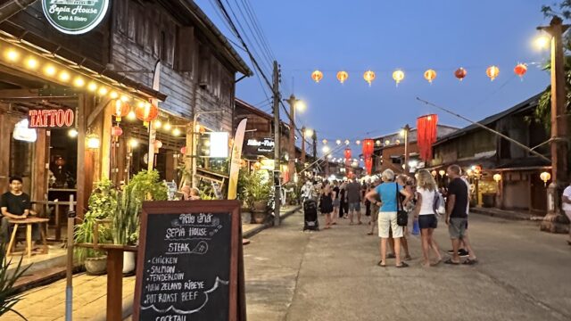 A lively street at dusk, with people walking, hanging lanterns, and a restaurant chalkboard menu in the foreground.
