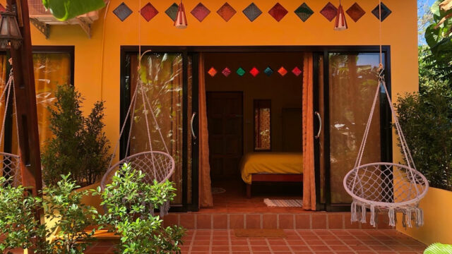 Yellow house with two hanging chairs, open glass doors, plants, and colorful diamond-shaped window accents above.
