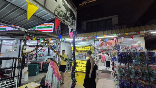 People walk through a brightly lit outdoor market decorated with colorful flags and tinsel at night.