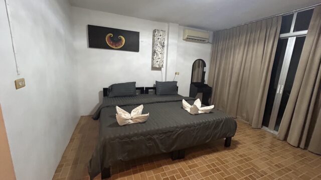 A neatly made bed with gray linens, towel swans, pillows, art above, and a small vanity in a room with tiled floor and curtains.