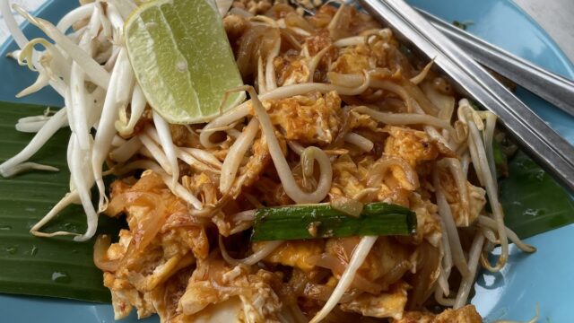 A plate of pad thai with tofu, bean sprouts, lime, chopsticks, and a small dish of sauce on the side.