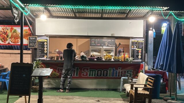 A person stands at an outdoor fruit smoothie stall at night, surrounded by chairs and colorful lights.