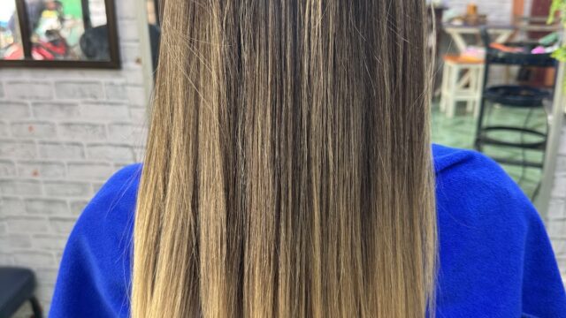 Woman with straight, long hair featuring dark roots and blonde ombre ends, seated in a salon chair, viewed from behind.