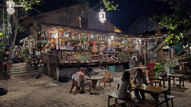People enjoy food and drinks at a lively, colorful beachside restaurant at night, with string lights and rustic decor.