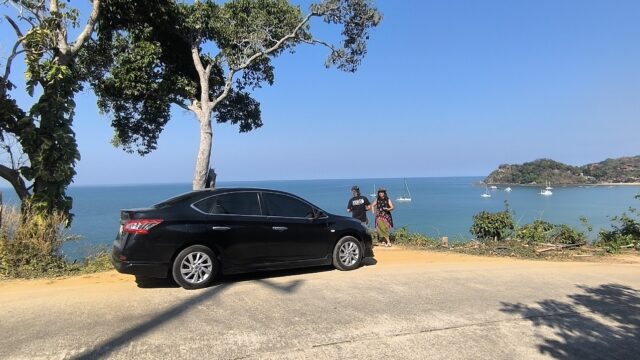 A black car parked by a coastal road with two people standing, overlooking the blue sea and boats.