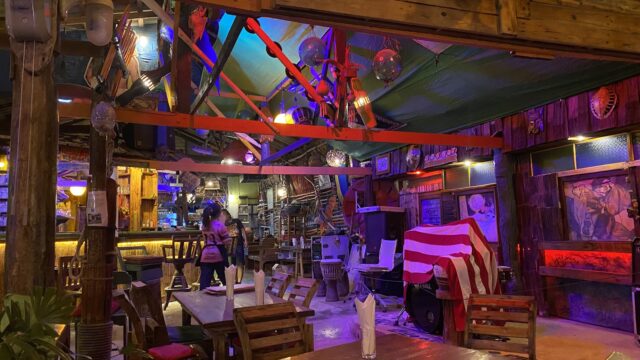 A cozy, colorful bar with wooden tables, eclectic decorations, dim lights, and a person walking near the counter.