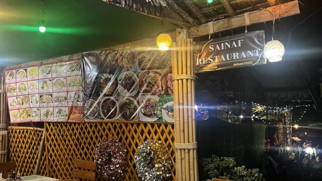Outdoor restaurant with wooden tables, menu photos on wall, hanging lights, and festive decorations under a thatched roof at night.