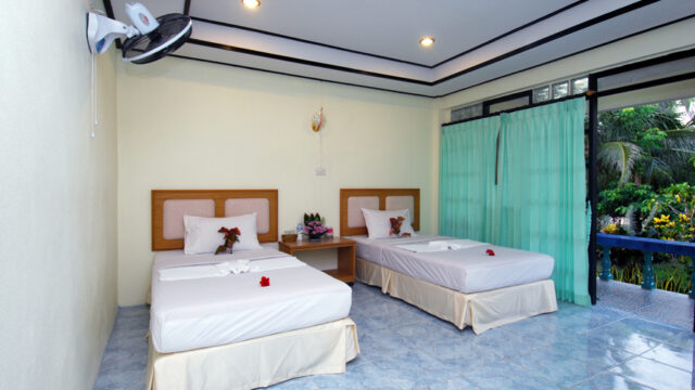 Hotel room with two single beds, flower decorations on the beds, green curtains, and a view of a garden through glass doors.