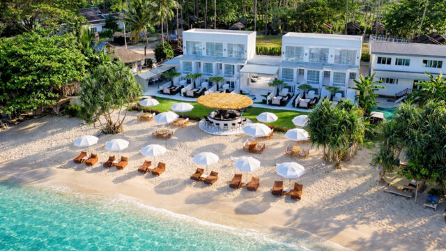 Aerial view of a beachfront resort with white umbrellas, lounge chairs, and turquoise water at the shoreline.